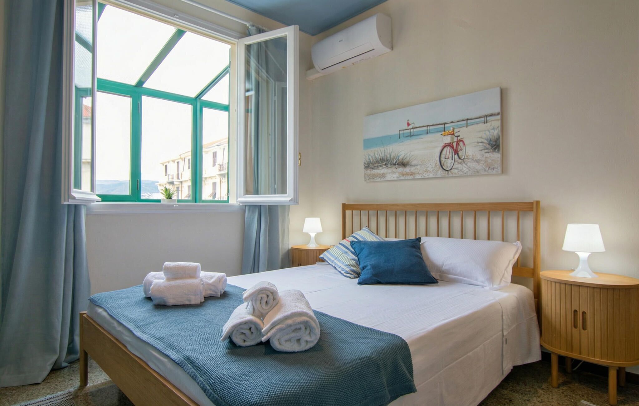 A bedroom with a bed, two bedside tables with lamps, a painting on the wall, and a window with curtains.
