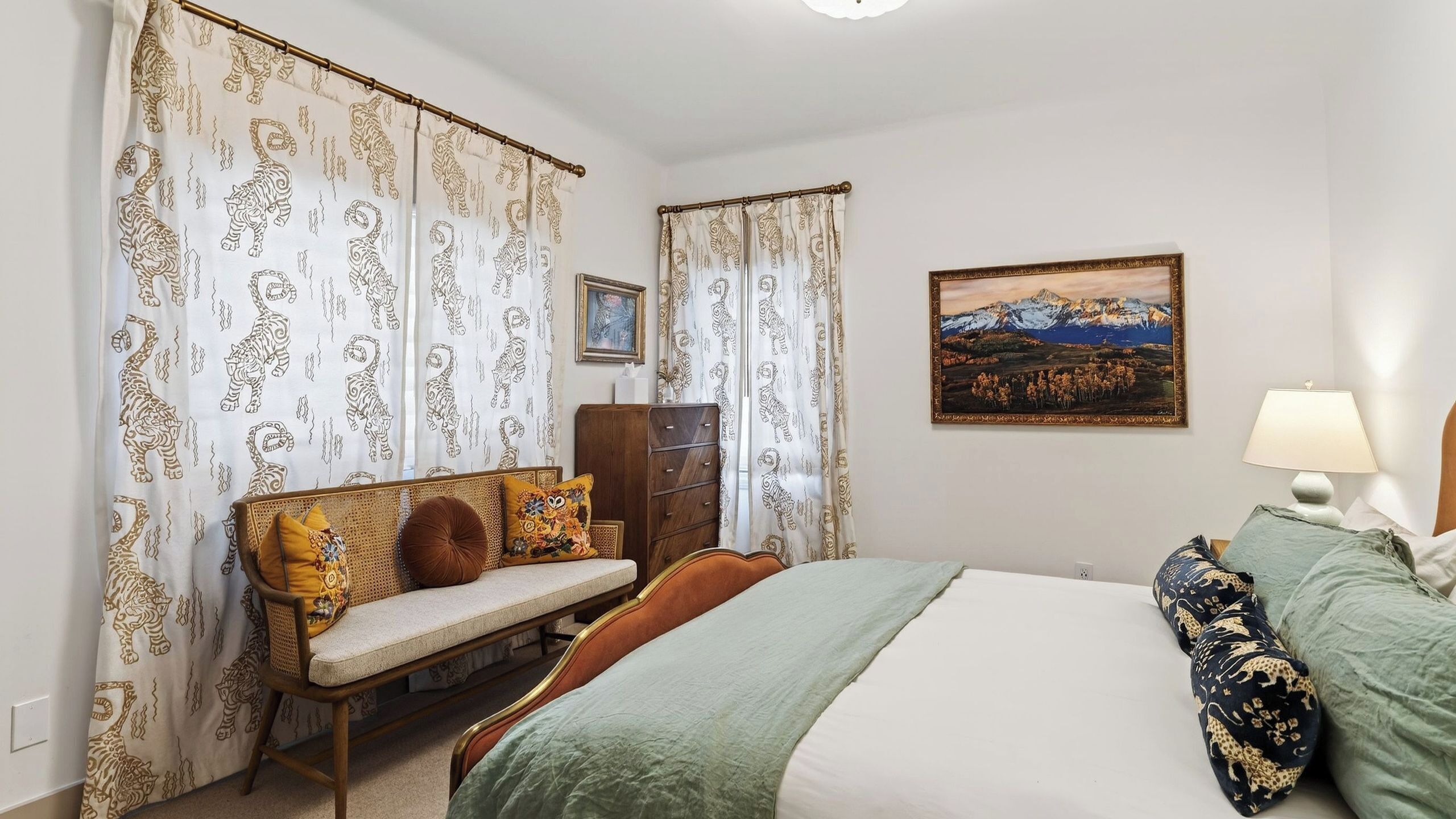 A bedroom with a bed, a bench with cushions, a dresser, a lamp, and a painting on the wall.
