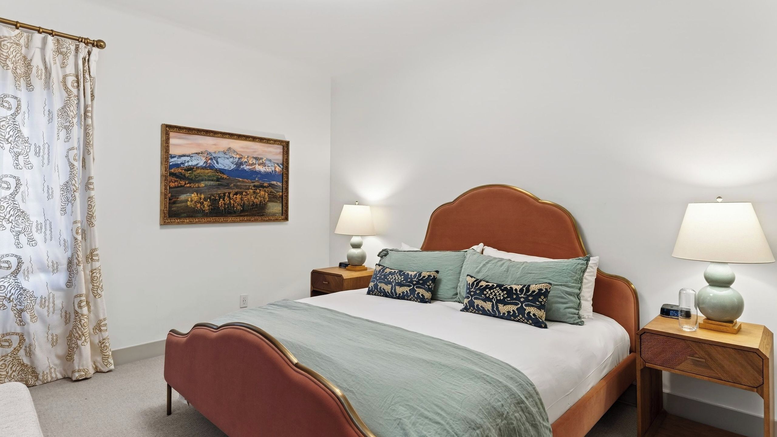 Une chambre à coucher comprenant un lit, deux tables de chevet, une commode, une lampe, un tableau et un rideau.