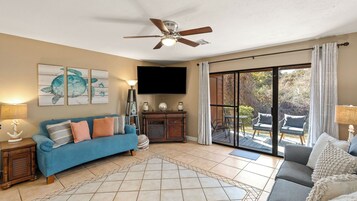 Ein Wohnzimmer mit blauem Sofa, grauem Sofa, Deckenventilator, Flachbildfernseher und Blick durch eine Glastür auf eine Außenpatio.