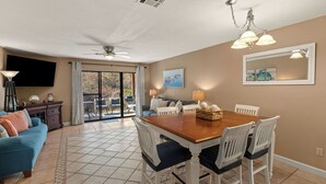 A dining area with a wooden table, white chairs, a ceiling fan, and a large window with a view of the outdoors. - Beachwalk 10B by Avantstay PCB Condo Across From the Beach + Pool & Tennis Court (Panama City Beach)