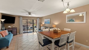 Comedor con mesa de madera, sillas blancas, ventilador de techo y ventana grande con vistas al exterior.