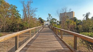 A wooden boardwalk with a metal railing, surrounded by trees and a building in the background. - Beachwalk 10B by Avantstay PCB Condo Across From the Beach + Pool & Tennis Court (Panama City Beach)