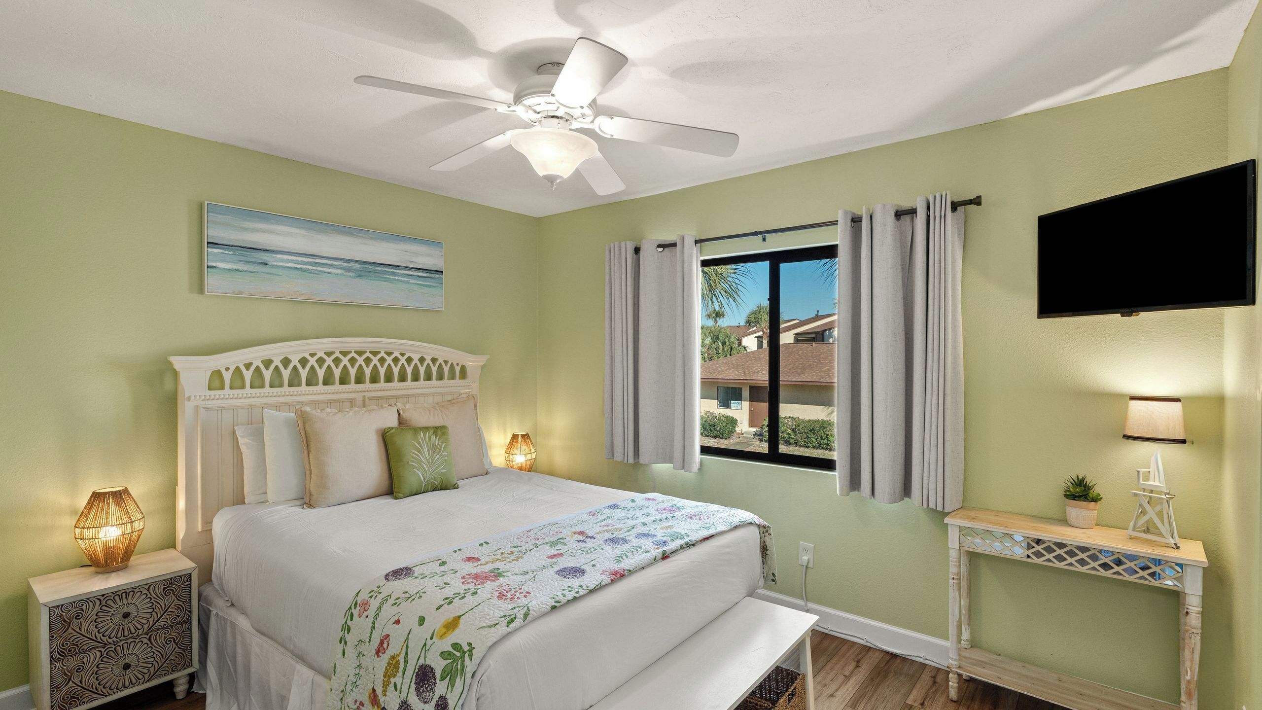 A bedroom with a bed, a ceiling fan, a window with curtains, a bedside lamp, a TV, and a small table with a plant.