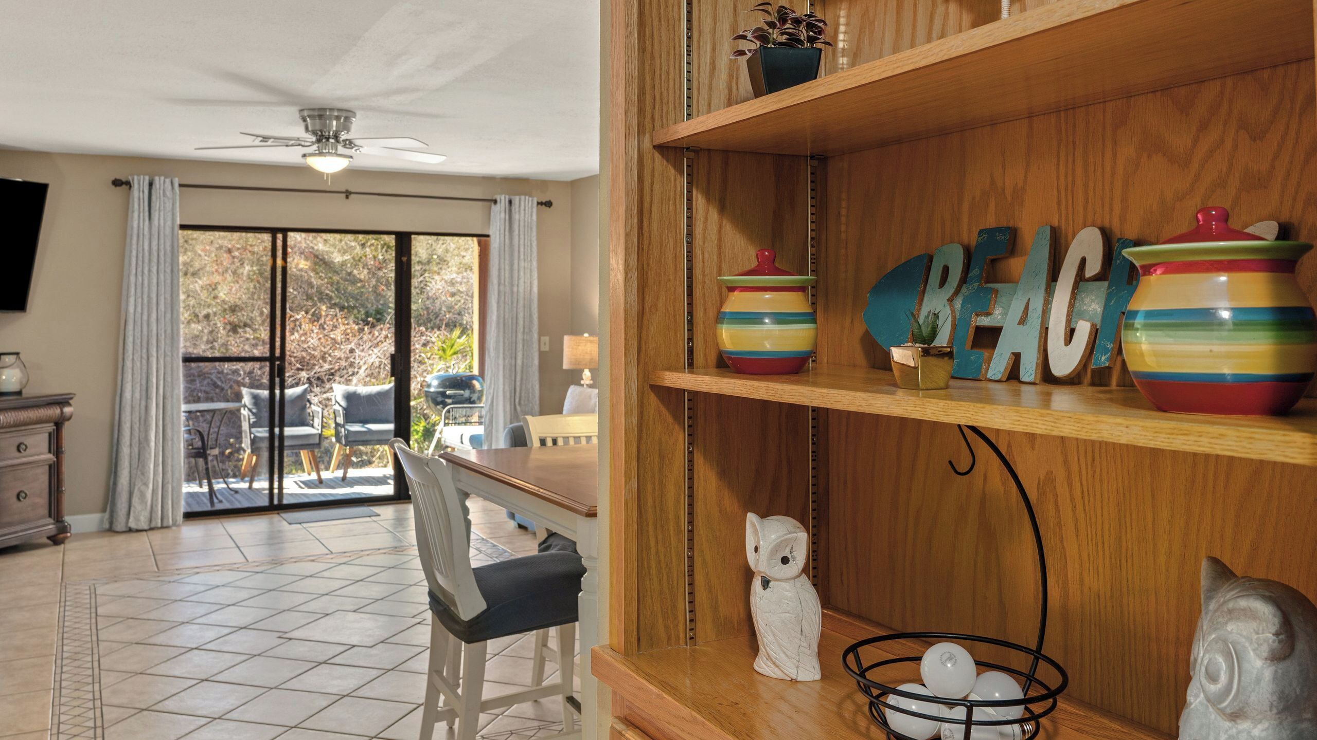 Quarto com área de refeições, uma prateleira de madeira com objetos decorativos e vista para um pátio exterior através de uma porta de vidro.