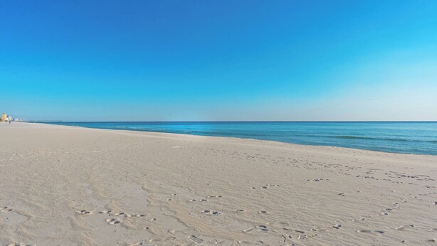 A wide sandy beach with footprints, a clear blue sky, and a distant pier. - Beachwalk 10B by Avantstay PCB Condo Across From the Beach + Pool & Tennis Court (Panama City Beach)