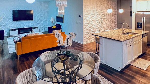 Een moderne keuken met een centraal eiland, een eetgedeelte met een glazen tafel, en een woonkamer met een bank en televisie.