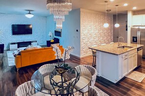 Een moderne keuken met een centraal eiland, een eetgedeelte met een glazen tafel, en een woonkamer met een bank en televisie.