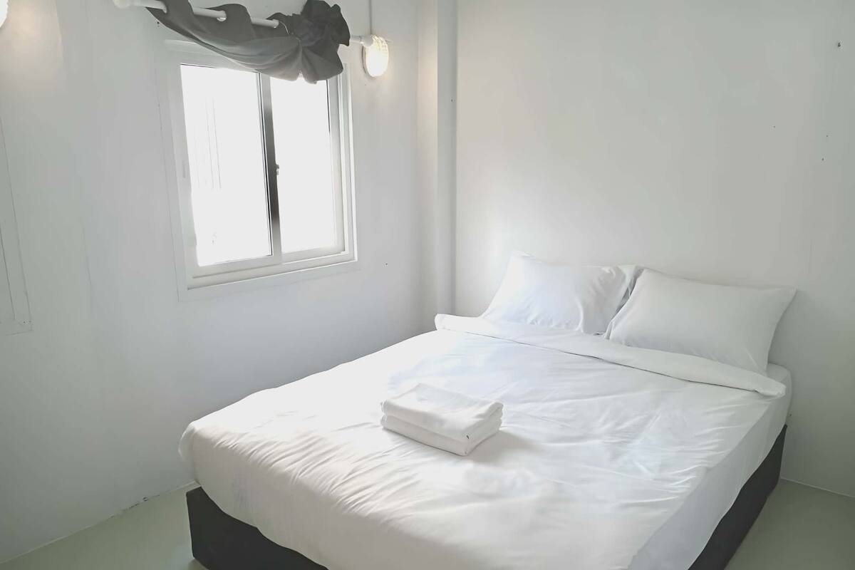 A neatly made bed with white linens and a folded towel, a window with curtains, and a wall-mounted light fixture.