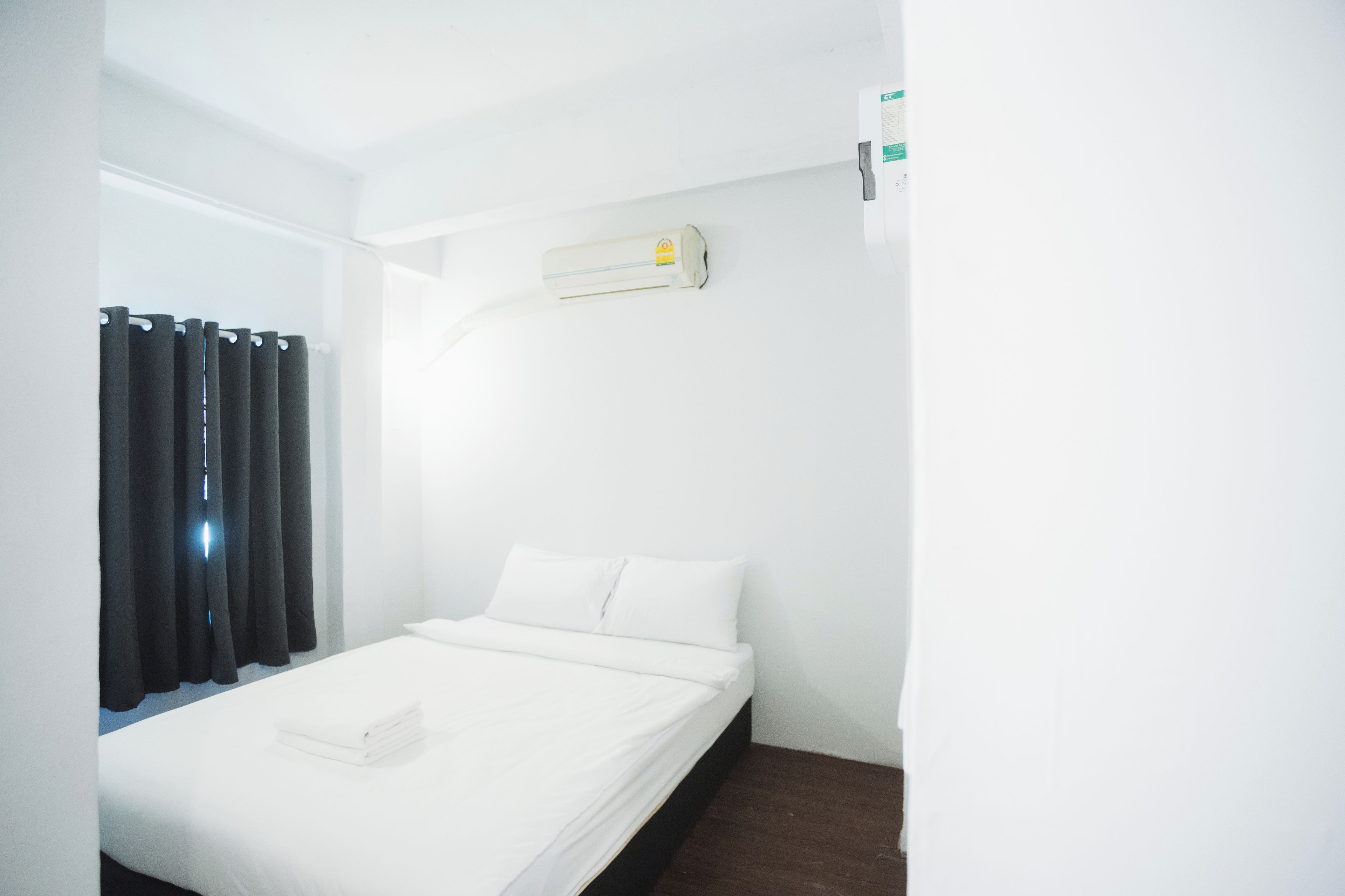 A single bed with white bedding, a dark wooden nightstand, a wall-mounted air conditioner, and black curtains on a white wall.