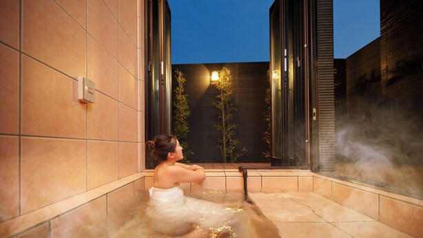 Une personne dans un bain à remous dont les murs sont recouverts de carreaux, avec vue sur des plantes de bambou à l’extérieur.