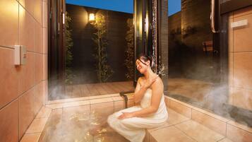 A person in a white bathrobe sitting in a steam room with tiled walls and a bamboo plant in the background.
