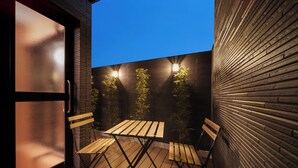 Une terrasse en bois avec une porte vitrée et un mur en briques. On y trouve deux tables et chaises en bois, ainsi que deux lampes fixées au mur.
