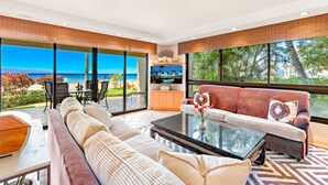 Ein geräumiges Wohnzimmer mit einer großen Couch, einem Couchtisch und einem Fernseher. Große Fenster bieten Blick auf Strand und Meer.