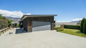 A modern garage with a large door, a paved driveway, and a well-maintained lawn with trimmed bushes. - Diamond on the Peninsula (Wānaka)