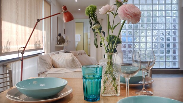 Un coin repas avec une table dressée pour deux personnes, agrémenté d’un bol bleu, d’un verre et d’un vase avec des fleurs.