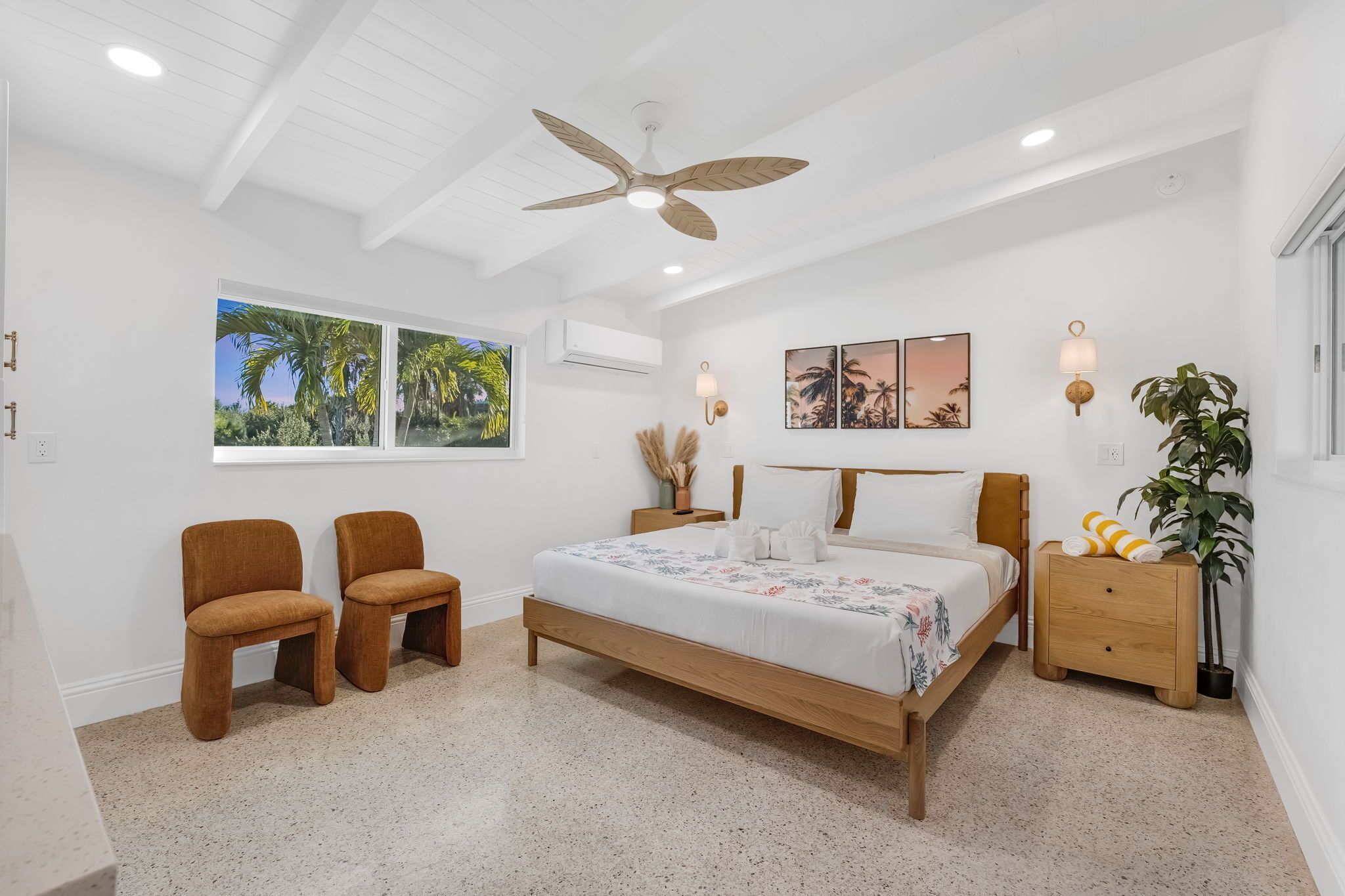 Quarto com cama, duas cadeiras, mesa de cabeceira, ventoinha no teto, luminárias fixadas na parede e quadros emoldurados.