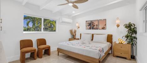 Quarto com cama, duas cadeiras, mesa de cabeceira, ventoinha no teto, luminárias fixadas na parede e quadros emoldurados.