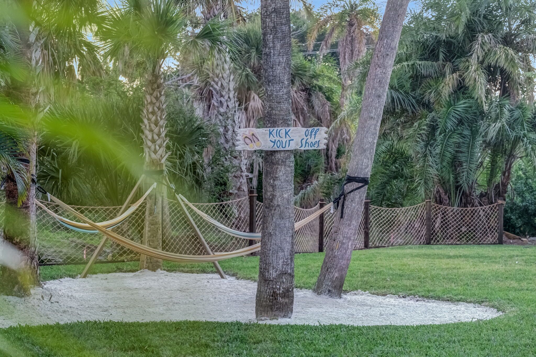 A hammock tied between palm trees, a sign that says "Kick off your shoes," and a fenced area with a sandy ground.