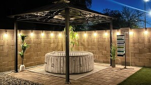 A covered outdoor seating area with a hot tub, surrounded by brick walls and illuminated by string lights. - Spacious 4BR w/ Backyard Spa Near Westgate Az (Youngtown)