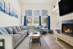 Una sala de estar moderna con un sofá seccional, una mesa de centro de madera, una chimenea y ventanas grandes con cortinas.
