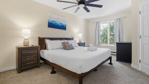 Une chambre à coucher comprenant un lit, des tables de chevet, un ventilateur de plafond, une fenêtre avec des rideaux et un tableau accroché au mur.