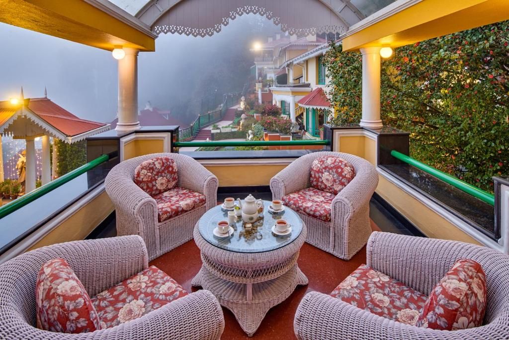 A covered outdoor seating area with wicker furniture and a central table, overlooking a garden and buildings.