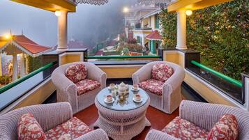 A covered outdoor seating area with wicker furniture and a central table, overlooking a garden and buildings.