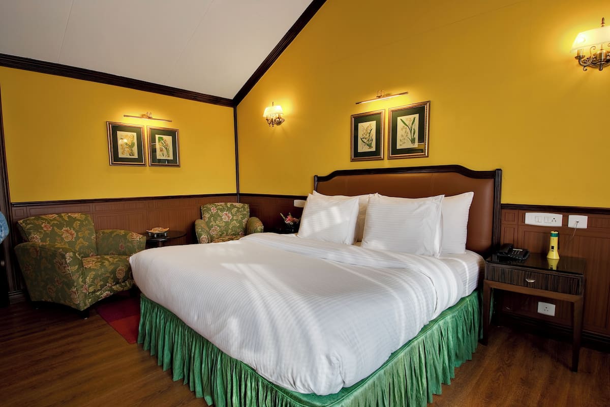 Een hotelkamer met een groot bed, een bedspreider met groene rok, bloemenarmstoelen en lijstwerken aan de muren.