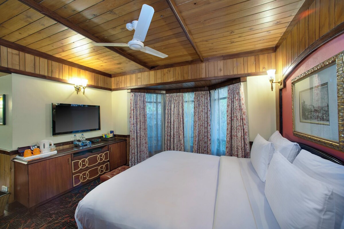 Quarto de hotel com uma cama grande, teto de madeira, televisão de tela plana, mesa com cafeteira e janela com cortinas florais.