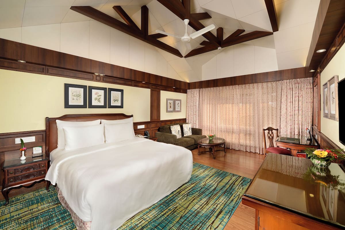 Een hotelkamer met een groot bed, een houten hoofdeinde, een bureau met een vaas, een stoel en een plafondventilator.