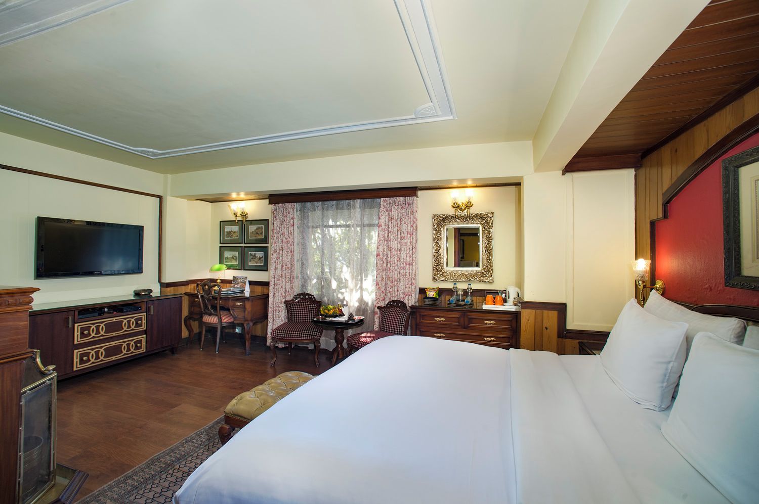 A hotel room with a large bed, a TV, a desk with a chair, a dining table with chairs, and a mirror.