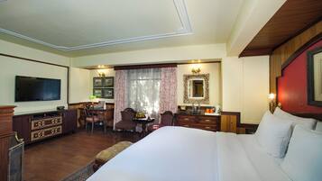 A hotel room with a large bed, a TV, a desk with a chair, a dining table with chairs, and a mirror.