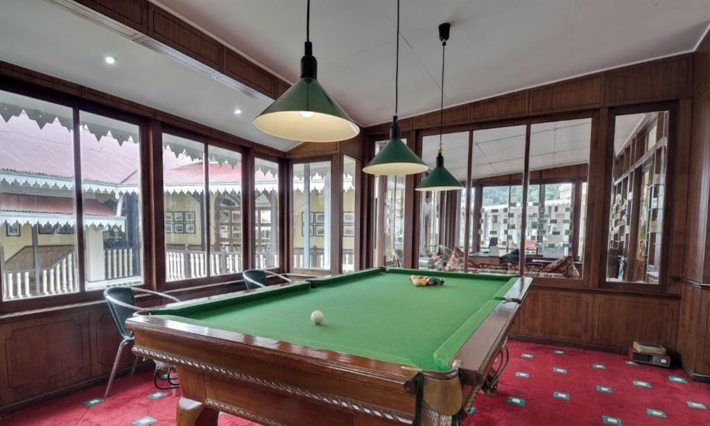 A pool table in a room with large windows, wooden paneled walls, and hanging pendant lights.