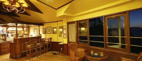 Zona de bar iluminada con muebles de madera y grandes ventanales con vistas a la noche.
