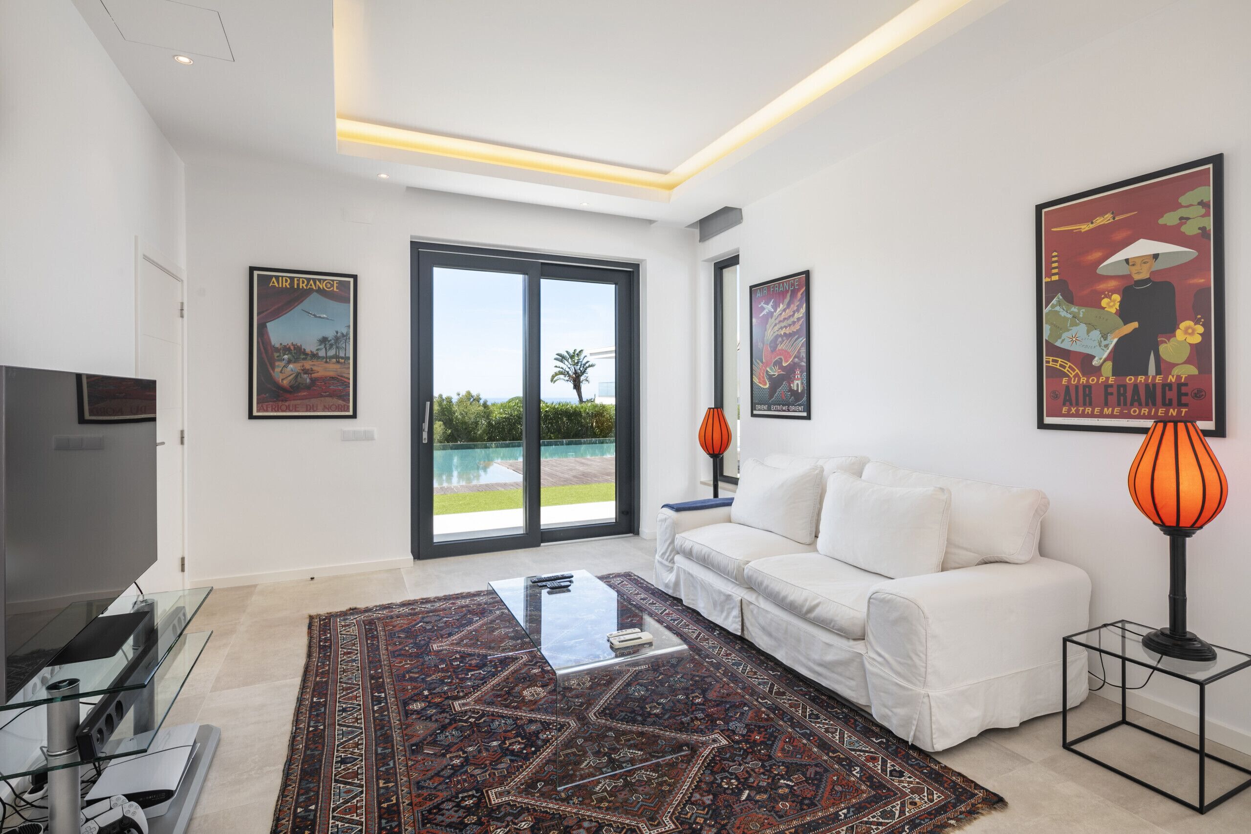 A modern living room with a white sofa, a glass coffee table, a patterned rug, and a large window with a view of a pool and greenery.