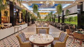 A spacious indoor dining area with round tables and wicker chairs, surrounded by high ceilings with hanging plants and a view of greenery outside. - Mayfair Tea Resort Siliguri (Siliguri)