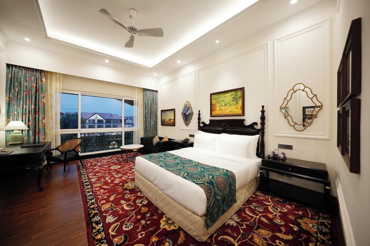 A hotel room with a large bed, a desk, a chair, a window with curtains, and a view of a building outside.