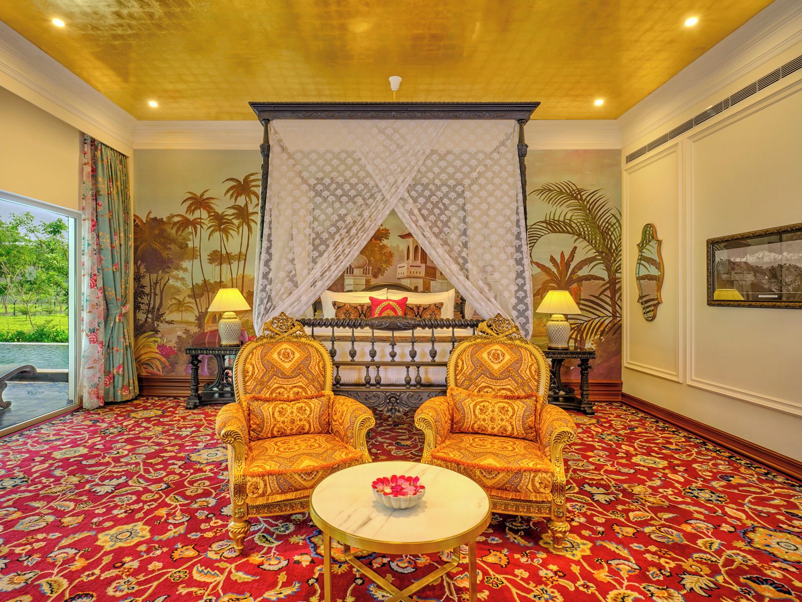 Une chambre avec un lit à baldaquin, deux fauteuils richement décorés, une table ronde ornée d’un vase de fleurs et un tapis rouge à motifs.