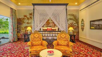Une chambre avec un lit à baldaquin, deux fauteuils richement décorés, une table ronde ornée d’un vase de fleurs et un tapis rouge à motifs.