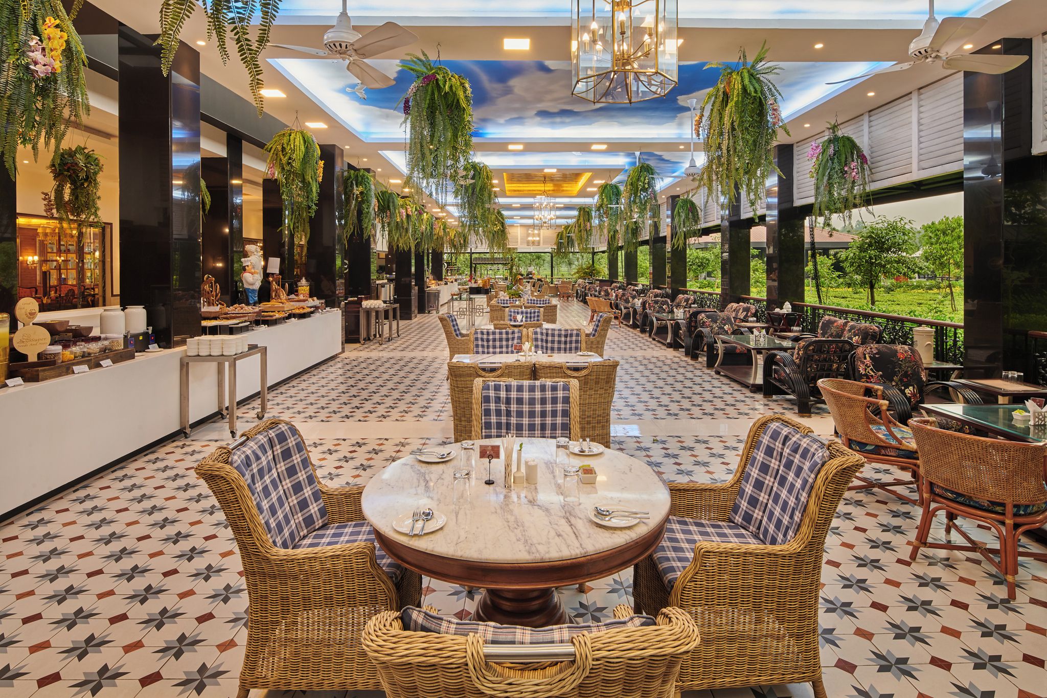 Un vaste espace de restauration intérieur, doté de tables rondes et de chaises en osier, agrémenté de hauts plafonds ornés de plantes suspendues et offrant une vue sur la verdure environnante.