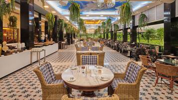 Un vaste espace de restauration intérieur, doté de tables rondes et de chaises en osier, agrémenté de hauts plafonds ornés de plantes suspendues et offrant une vue sur la verdure environnante.