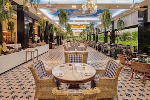A spacious indoor dining area with round tables and wicker chairs, surrounded by high ceilings with hanging plants and a view of greenery outside. - Mayfair Tea Resort Siliguri (Siliguri)