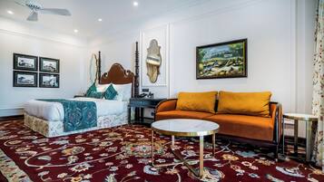 Una habitación con una cama, un sofá, una mesa de centro redonda, un ventilador de techo, cuadros enmarcados y un cuadro en la pared.