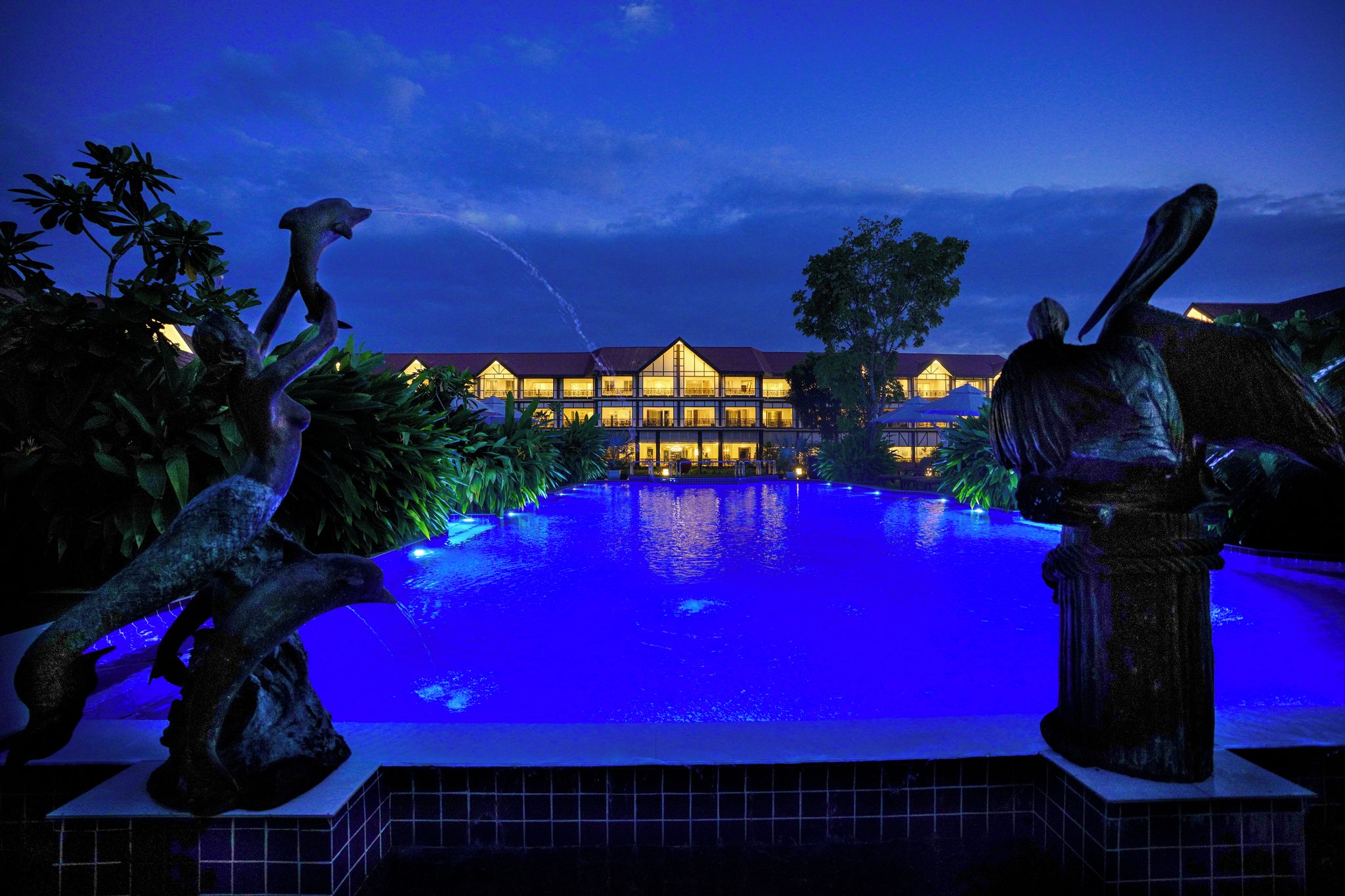 Vista nocturna de un hotel con piscina, iluminado con luces azules y rodeado de estatuas.