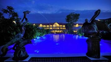 Vista nocturna de un hotel con piscina, iluminado con luces azules y rodeado de estatuas.