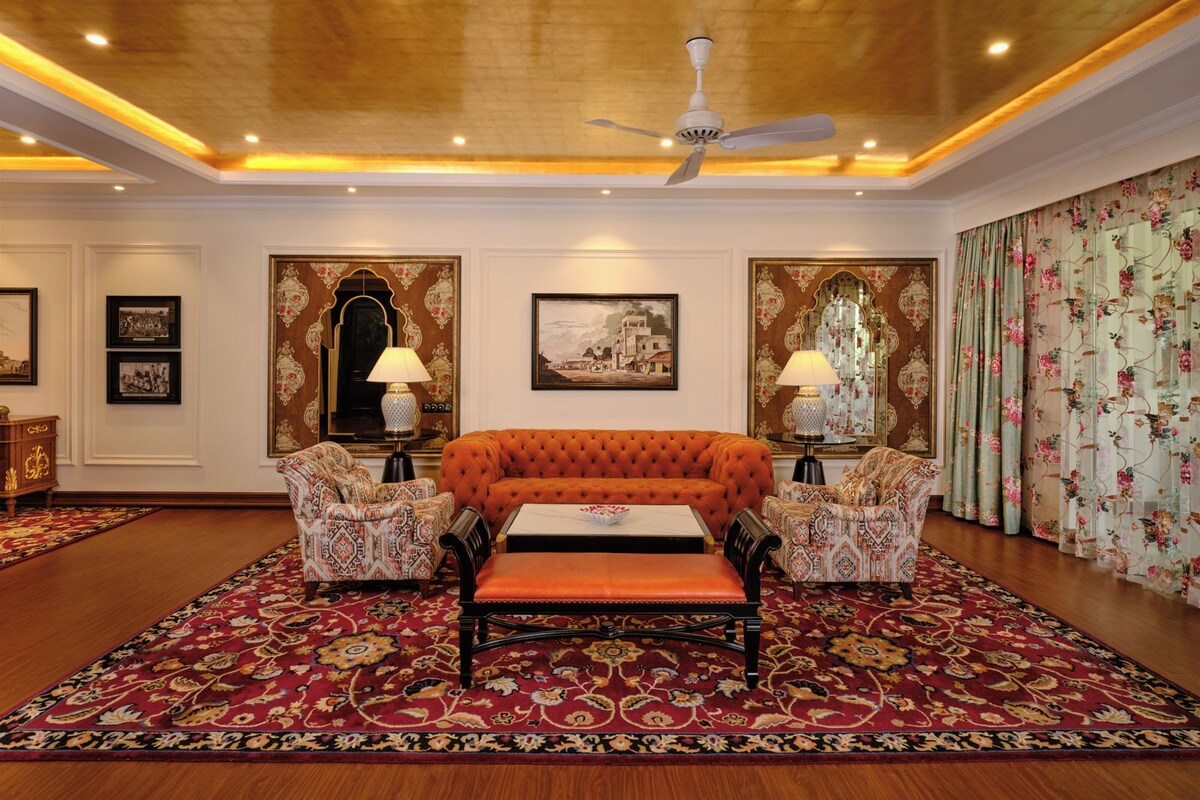 Una habitación con un sofá capitonado, sillones estampados, alfombra roja y decoraciones ornamentales en las paredes.