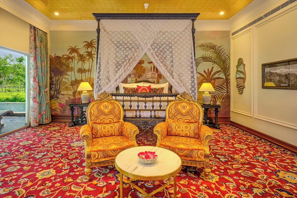 A room with a canopy bed, two ornate chairs, a round table with a flower vase, and a red patterned carpet.