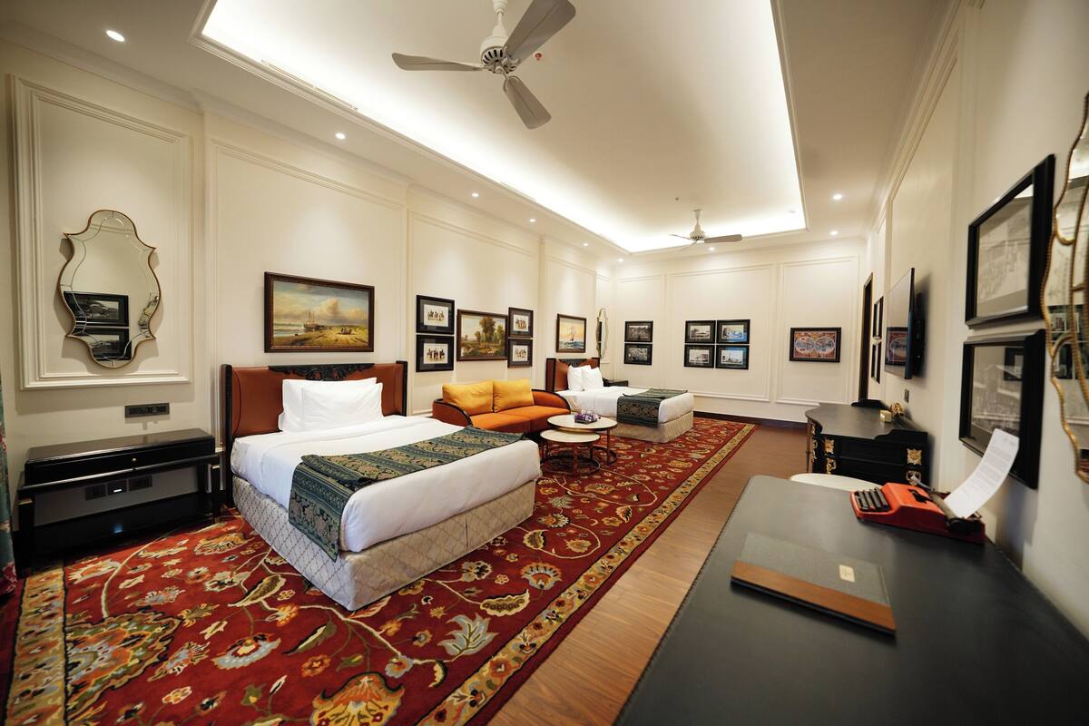 A hotel room with a large bed, a sofa, a desk with a typewriter, and a red carpet.