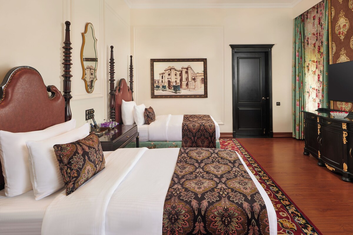 Habitación de hotel con dos camas, un espejo grande, un cuadro en la pared y un televisor sobre un mueble.